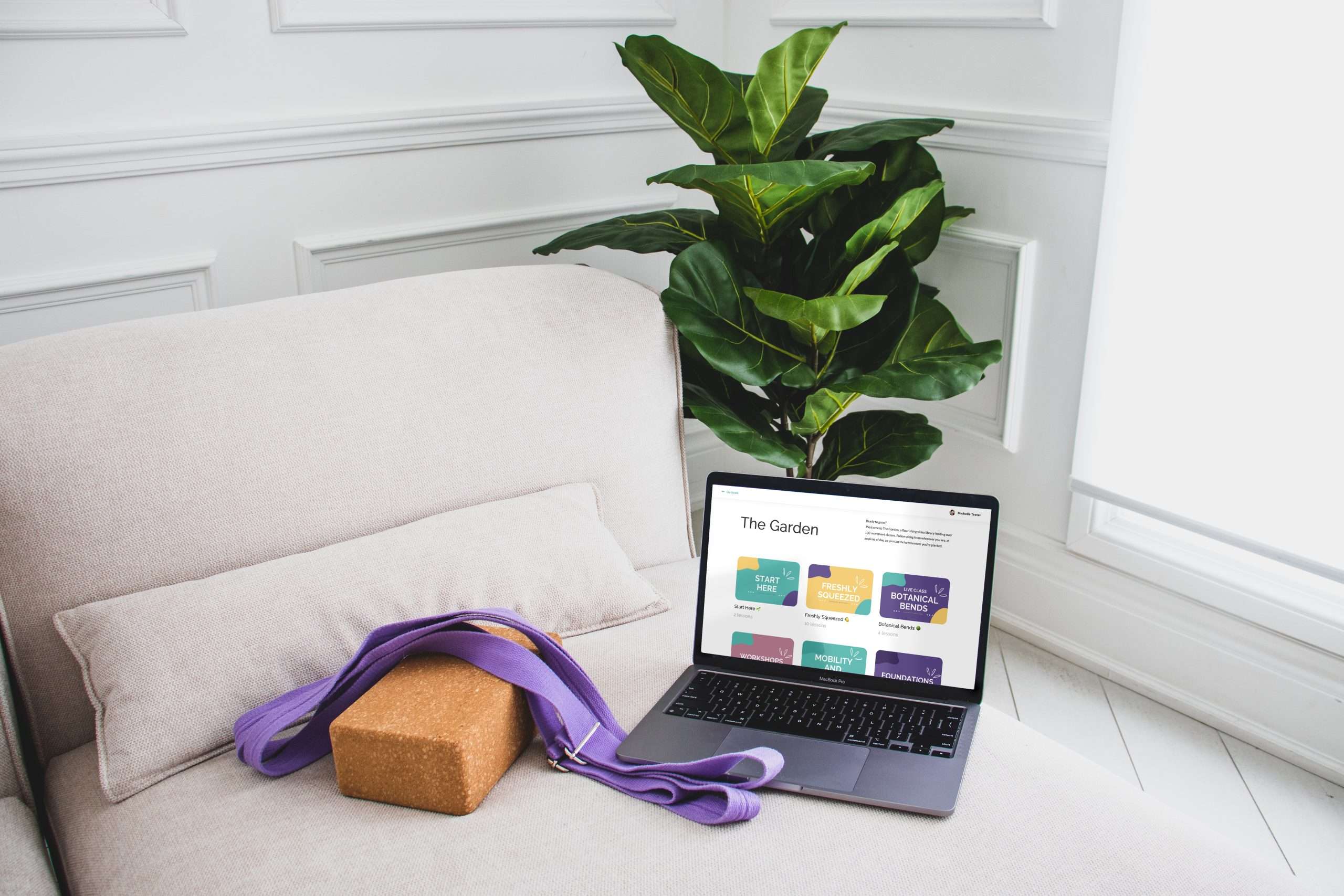 Macbook sitting on white couch, beside a cork yoga block with a purple yoga strap drapped over top. The macbook shows an image of The Garden, the on-demand library. Behind the couch is a green plant.