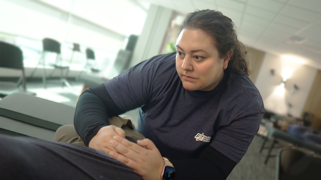 Vera is wearing a navy shirt that says Fascial Stretch Therapy on it. She has her hands cupped around the side of the hips on her client, looking intently off towards the client's shoulder.
