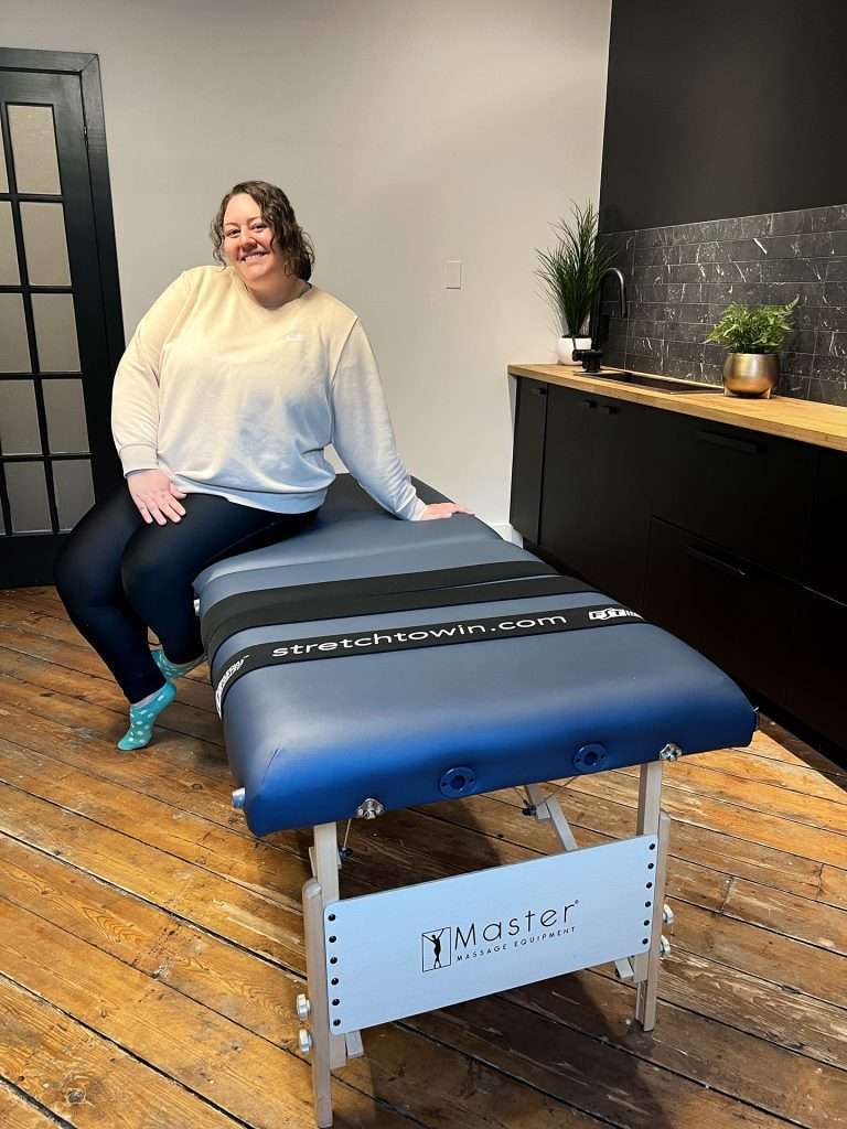 Vera sitting on the edge of a massage table where fascial stretch therapy in toronto happens. At the end of the massage table are three straps, one says "Stretch to Win" with the Fascia Stretch Therapy logo on it